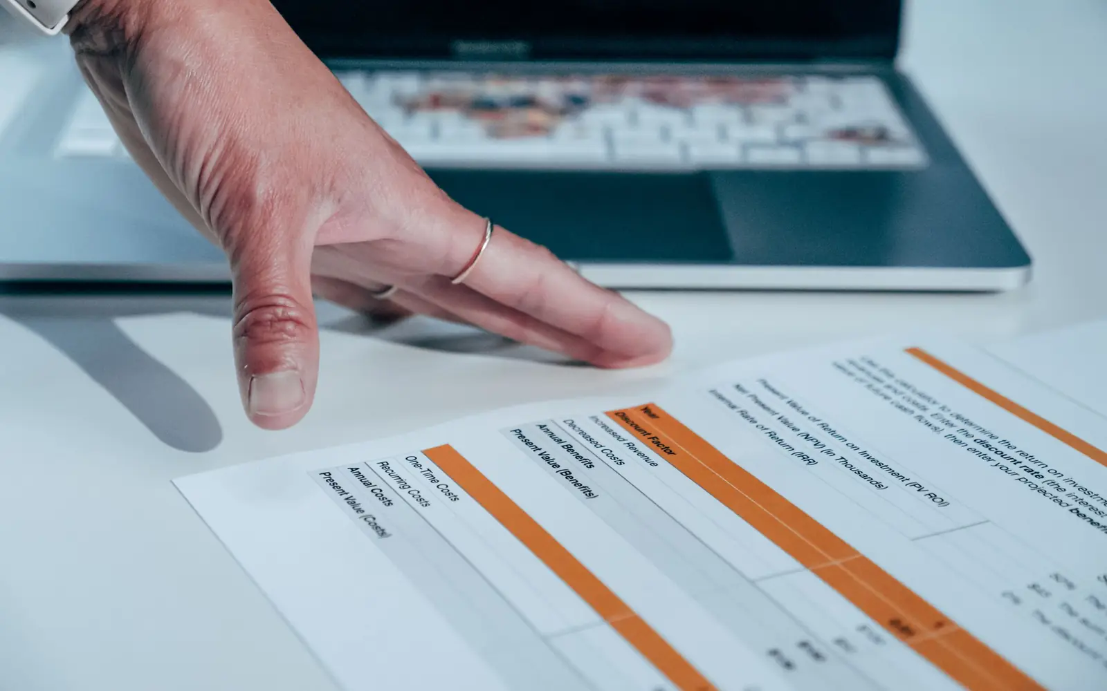 A hand pointing at a printed worksheet of figures on a desk, beside an open laptop.