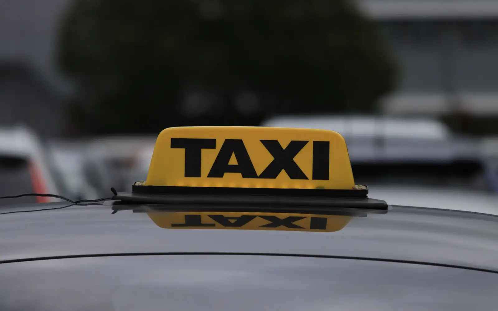 A lit yellow TAXI roof sign on the roof of a dark car, with a street out of focus behind it.