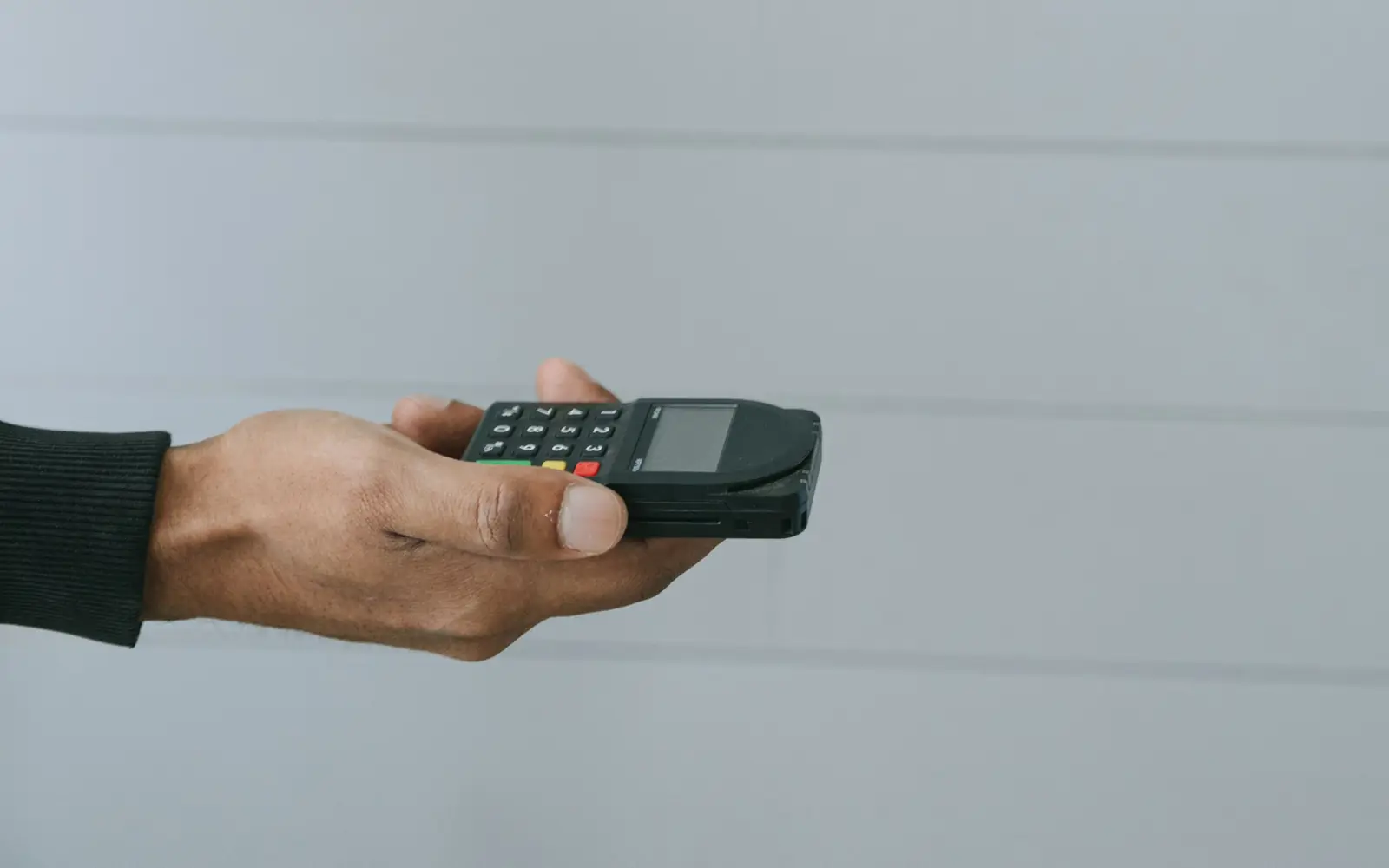 A hand holding a small card payment terminal with a numeric keypad, against a plain wall.